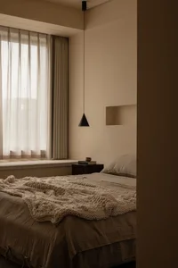 A minimalist bedroom with neutral-toned fabrics, featuring a bed, window with curtains, and a hanging lamp.