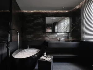 A minimalist modern bathroom featuring black tiles and concrete surfaces with a freestanding bathtub filled with bubbles unde