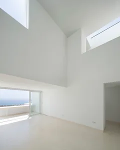 An empty minimalist living room with high ceilings, large glass windows overlooking the ocean, and white walls and floor.