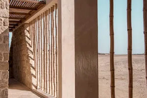 A wooden frame with stone walls extends from a desert-rimmed beach, showcasing the structure of a house.