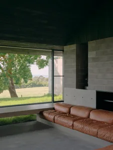 A modern living room featuring concrete walls and a leather sofa with a large window overlooking a grassy field.