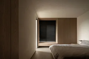 Minimalist bedroom featuring a bed with black blinds and wooden furniture against a white wall and hardwood floor.