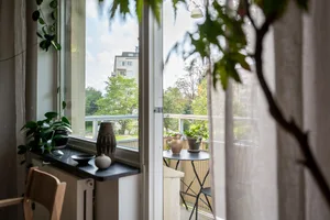 A minimalist apartment balcony with a glass and metal railing, a black table with a pitcher and vase, and a curtain with a vi