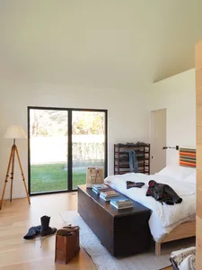 A modern minimalist bedroom featuring a wooden bed frame and a large window with a view of the grassy yard. The room is beaut