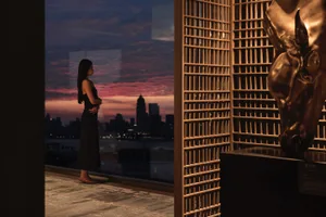 A woman standing on a wooden floor with a steel railing, observing a cityscape at sunset through a large window.