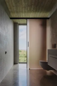 Modern industrial-style bathroom with concrete floor and wood paneling, featuring a frosted glass sliding door and a window o