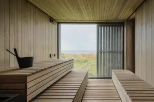 Modern industrial-style sauna with wooden benches and a large window overlooking a grassy field.
