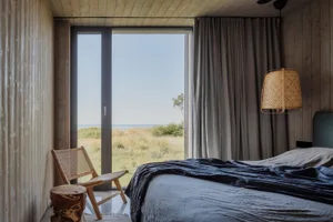 A serene minimalist bedroom with a large window overlooking a natural landscape, featuring a wooden bed, chair, and stool, an