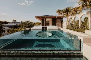 Modern swimming pool patio featuring a circular water feature, lounge chairs, and tropical landscaping.
