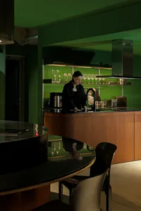 A modern kitchen with green walls, wooden cabinets, and glass shelves.