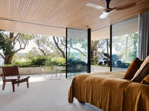 Modern bedroom with wooden ceiling and floor, featuring a large bed, a chair, and a view of trees and a garden.