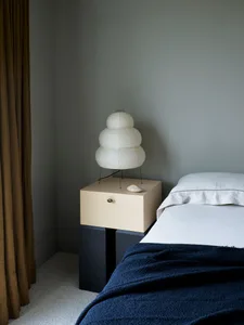 A minimalist bedroom with a bed, a wooden nightstand, a white and beige light fixture, and a navy blue knit blanket.