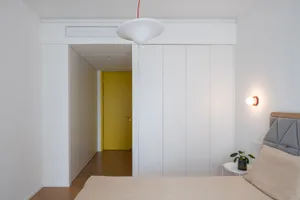 Modern minimalist bedroom interior with white walls, a white pendant light, and a yellow door in the background.