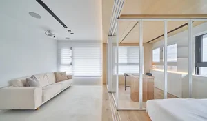 A modern living room with a neutral color palette featuring a comfortable sofa, blinds, and a study area with a desk and wind