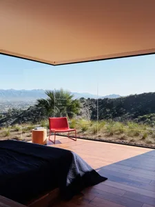 A modern residential bedroom with a wooden floor, a red chair, and a view of the mountains through a large glass window.