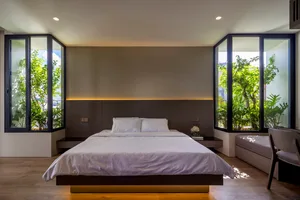 Modern minimalist bedroom featuring a sleek white bed with natural light from large windows overlooking greenery.