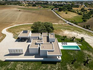 Aerial view of a modern villa with concrete finishes, featuring a swimming pool and a carport in a rural area.