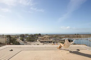 A person sitting in a white chair on a balcony with a metal railing overlooking a coastal landscape.