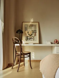 Minimalist bedroom with a wooden chair, a desk with a framed abstract painting, and a bed covered in beige textiles.