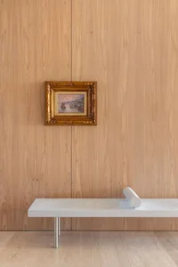 A minimalist interior design featuring a wooden frame with a framed painting and a modern, sleek bench with a cylindrical obj