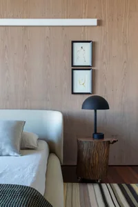A modern minimalist bedroom featuring a white bed with pillows, a wooden side table, a black table lamp, and two framed art p
