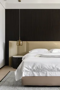 A modern minimalist bedroom with a beige headboard, white bedding, and a dark wood accent wall.