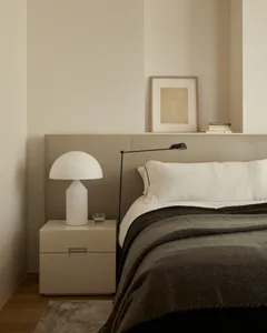 A minimalist bedroom with a white bed, a nightstand with a white lamp, and a floating shelf above the bed.