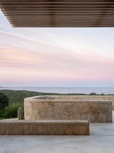 A minimalist architectural exterior featuring granite walls and a wooden louvered roof, overlooking the sea and sunset.