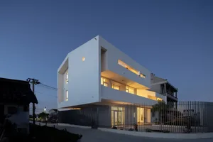 A modern, white residential building with warm interior lighting against a dark, twilight sky.
