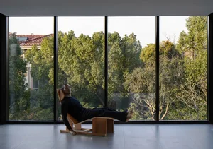 A man lying on a wooden relaxation chair in front of large windows with a view of trees.