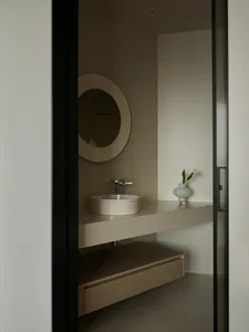 Modern minimalist bathroom with black framed glass door, white floating vanity, white sink, and white vase with green plant.