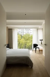 Bedroom with minimalist design, featuring a bed with wood headboard and a chair, natural light from large windows, and wooden