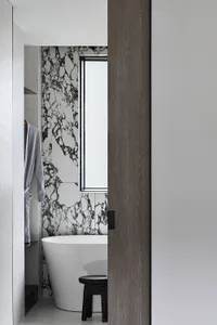 A minimalist bathroom featuring a white bathtub with a marble accent wall, a wooden stool, and a grey robe hanging