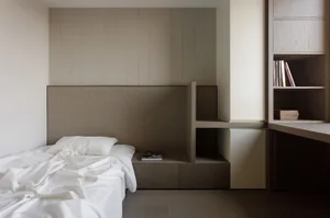 Minimalist bedroom with wooden furniture and white bedding, featuring integrated shelving and storage systems.