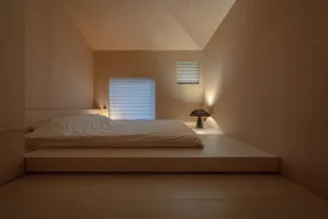 A minimalist bedroom with a wood floor, bed, and window with blinds.
