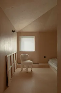 A minimalist bedroom with brown plywood walls, a window with blinds, and a small white chair.