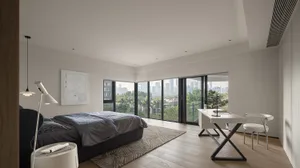 A modern minimalist bedroom featuring a large bed with dark bedding, a white desk, and floor-to-ceiling windows overlooking a