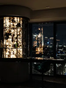 A modern bar counter featuring glass shelves and lighting, with a view of a cityscape at night.