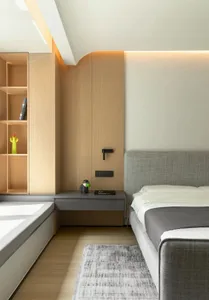 A modern bedroom featuring a grey upholstered bed with a floating nightstand and wooden shelving unit, illuminated by soft am