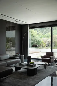 Modern living room featuring leather and glass furniture with a metal floor lamp and large windows overlooking greenery.