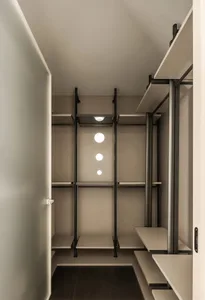 A minimalist walk-in closet featuring a black metal framework with white shelves and two circular lights mounted on the ceili