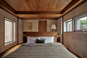 A modern bedroom featuring wooden walls, ceiling, and headboard, with white bedding and a lamp on the nightstand.