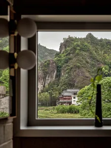 A window overlooking a lush mountain landscape with a traditional building nestled among the trees.