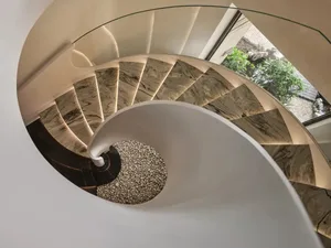 A modern spiral staircase featuring marble steps and glass railings with a white wall and green foliage in the background.