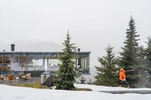 Modern residential building with glass walls and steel accents surrounded by snow and evergreen trees, with a person skiing i