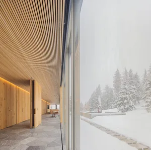 Modern minimalist interior with wood slat ceiling, stone flooring, and a large glass window overlooking a snowy forest.