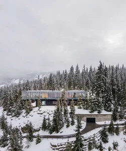 Modern glass lodge on snowy mountainside surrounded by evergreen trees.