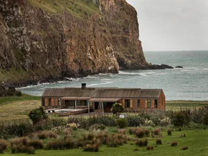 A modern, wooden house with a metal roof on a grassy hillside overlooking the ocean and a rocky cliff.