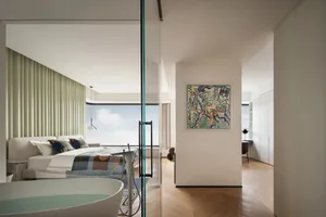 A minimalist bedroom with a large window, a queen-sized wooden bed with a patterned bedspread, and a glass bathtub.