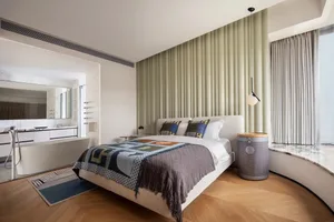A modern minimalist bedroom with a light wooden floor, white walls, and a large bed with geometric patterned bedding.